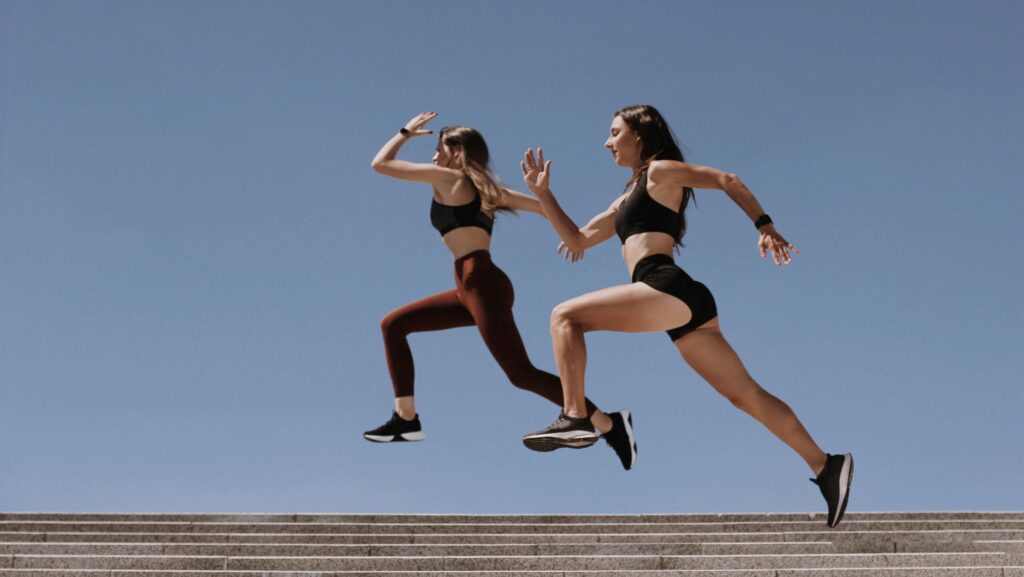 Two women in active wear leap energetically on stairs against a clear blue sky.